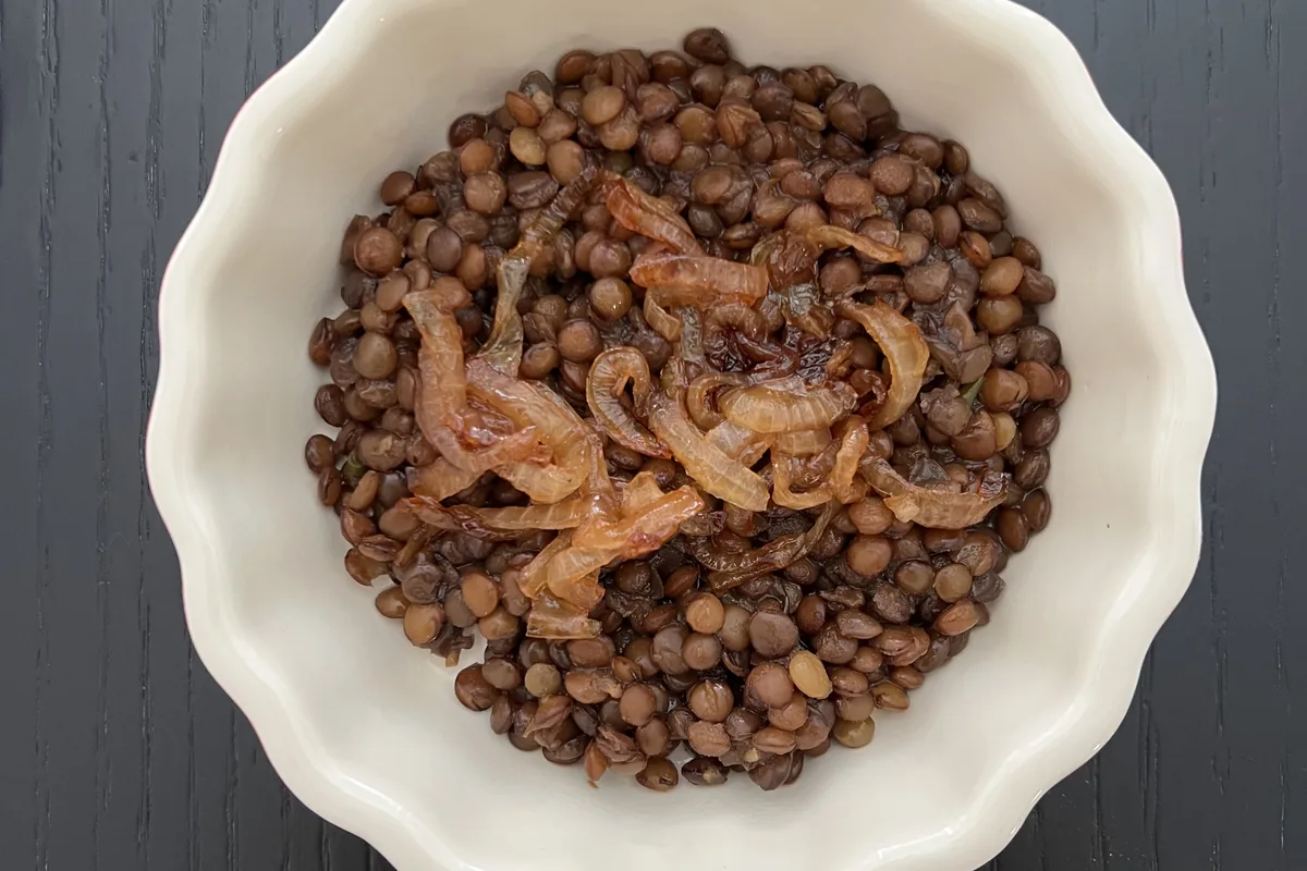 A white bowl of cooked brown lentils topped with caramelized onions