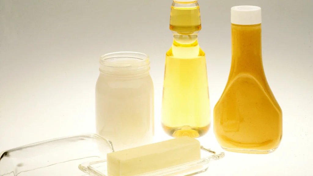A stick of butter on a glass dish beside a jar of mayonnaise, a bottle of oil and a bottle of salad dressing
