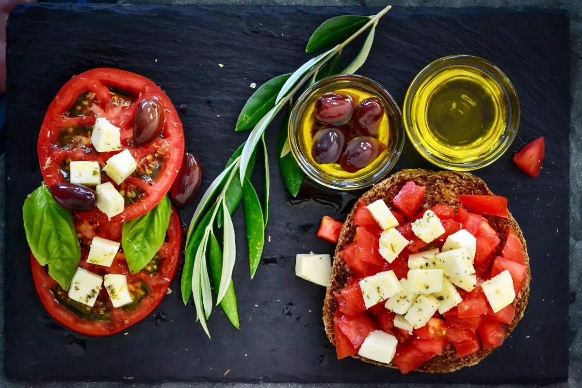 A barley rusk topped with diced tomato and feta cubes, with olives and olive oil on a slate board