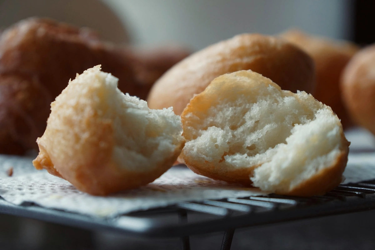Golden fried dough balls cooling on a wire rack, one broken open to show the airy inside