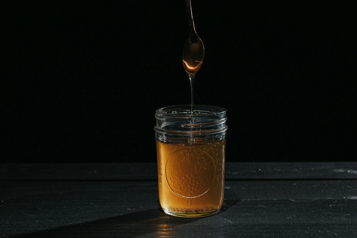 Honey dripping from a dipper into a small glass jar against a dark background