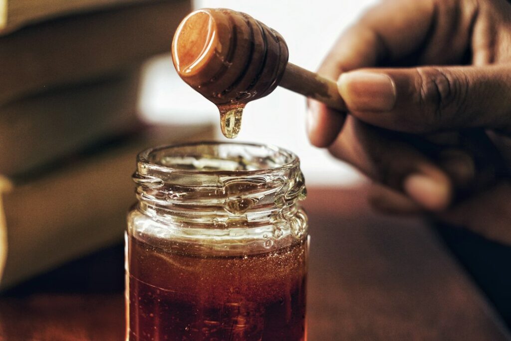 Golden honey dripping from a wooden dipper into a glass jar