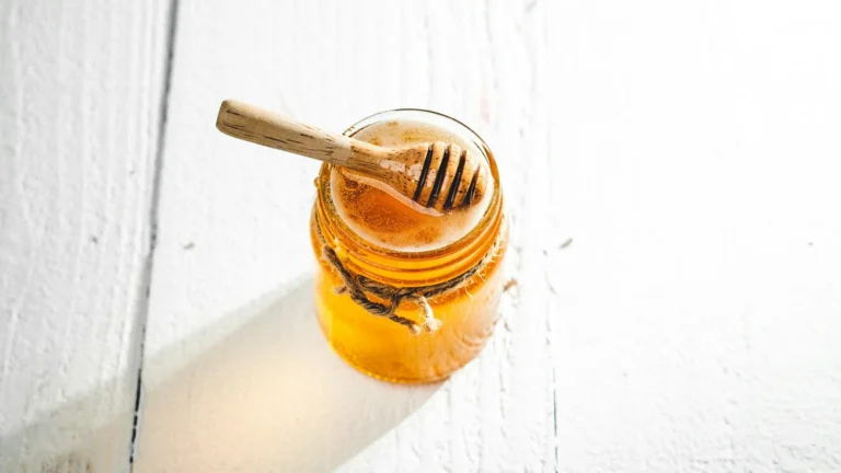 Jar of golden honey with a wooden dipper on a white wooden table, for reading honey nutrition facts