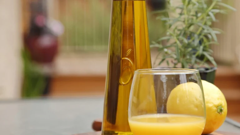 A glass bottle of olive oil with lemons and a glass of juice on a kitchen table