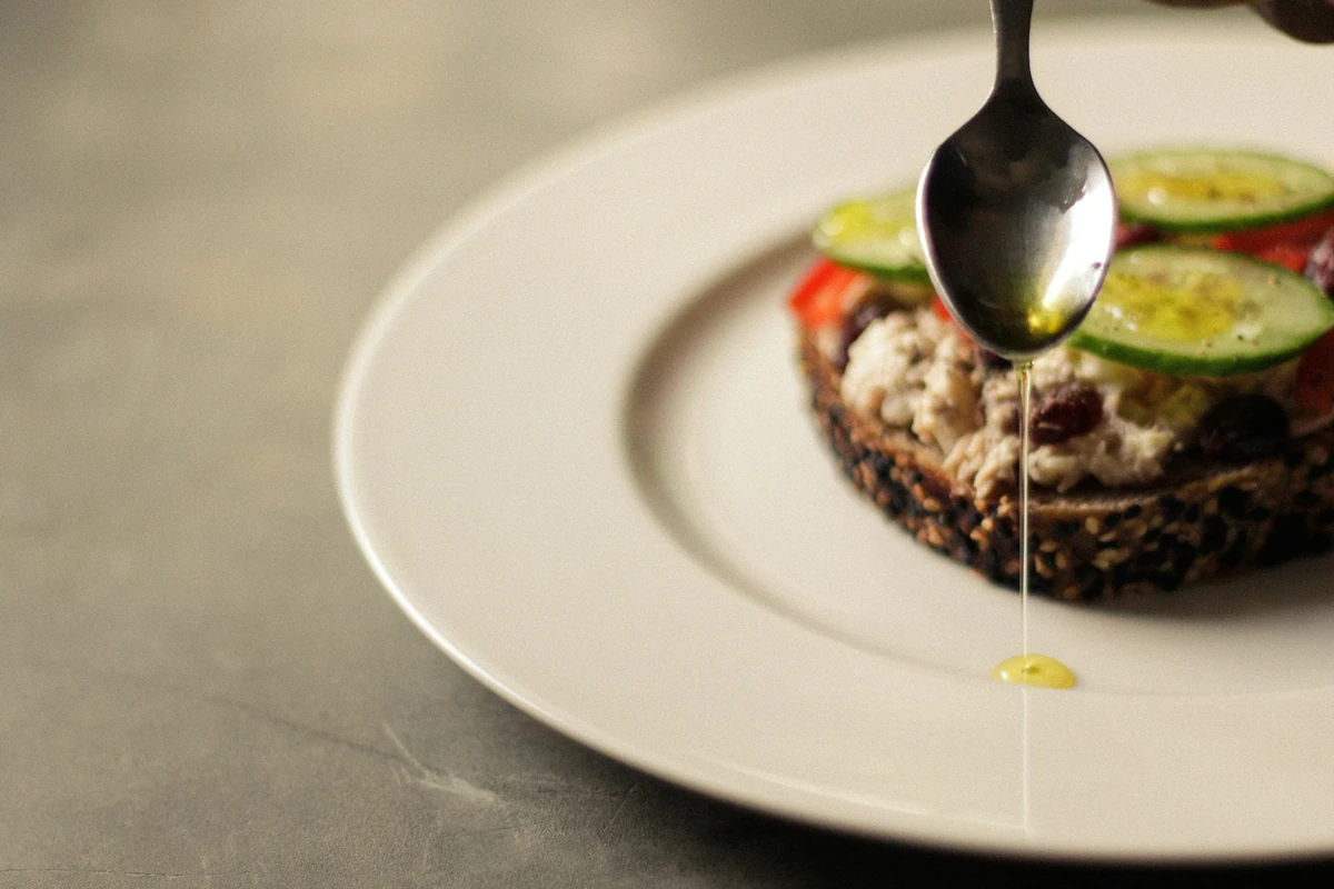 Spoon drizzling a thin stream of oil beside an open sandwich topped with cucumber