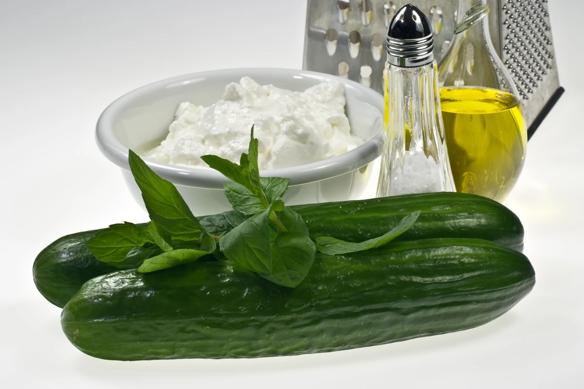 A bowl of tzatziki beside whole cucumbers, mint and a bottle of olive oil